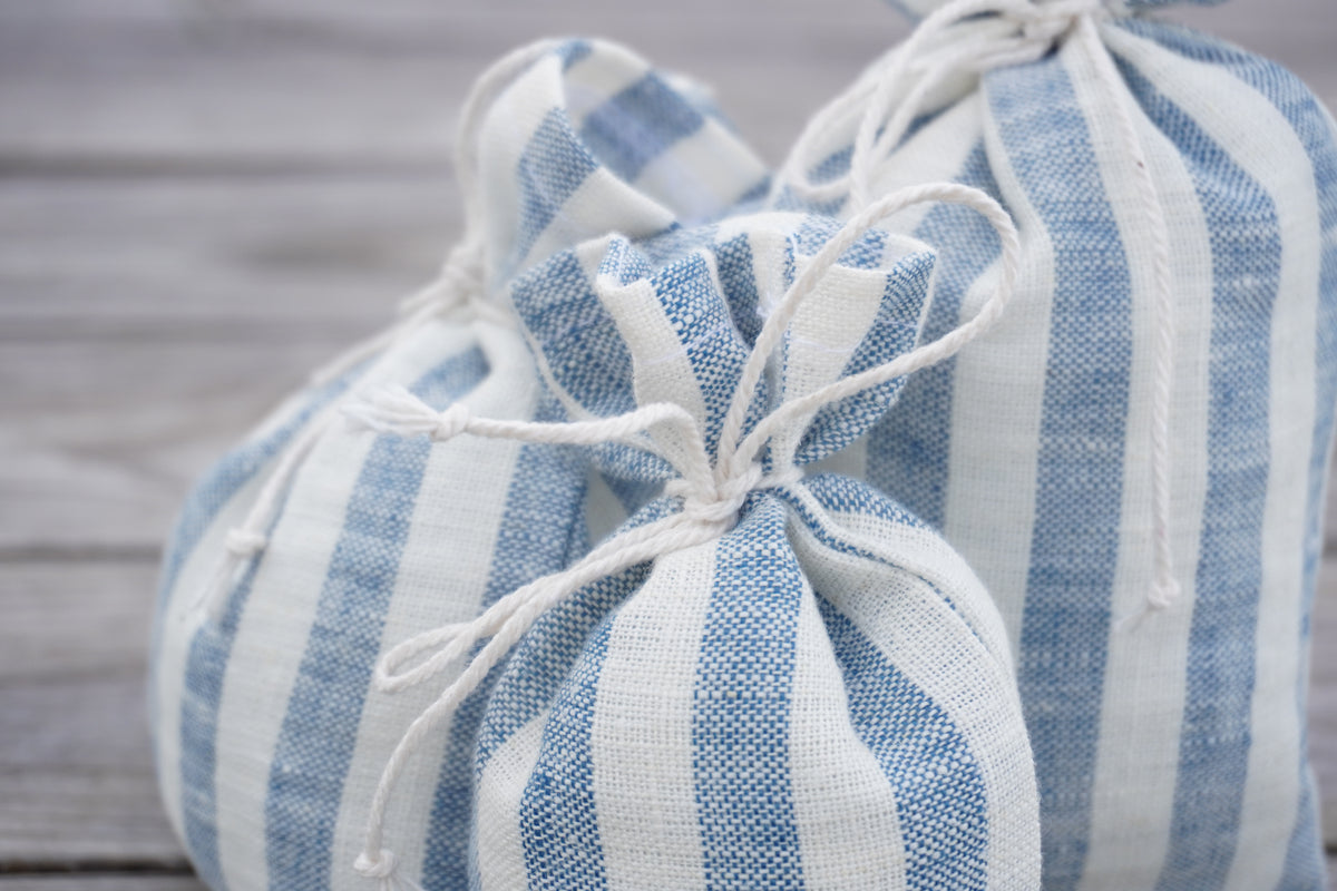 Two small gift bags made of blue and white striped linen fabric, tied with white cord, resting on a light gray wooden surface.