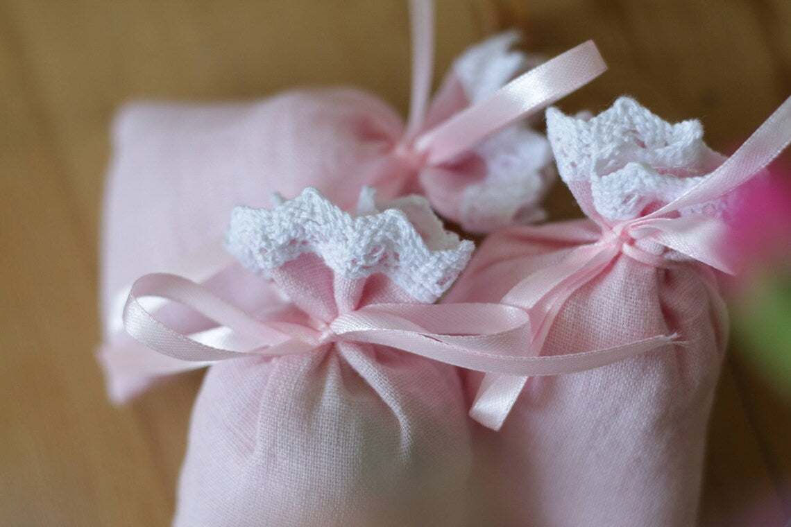 Three small pink linen  fabric bags tied with pale pink ribbons and decorated with white lace trim, arranged on a wooden surface.