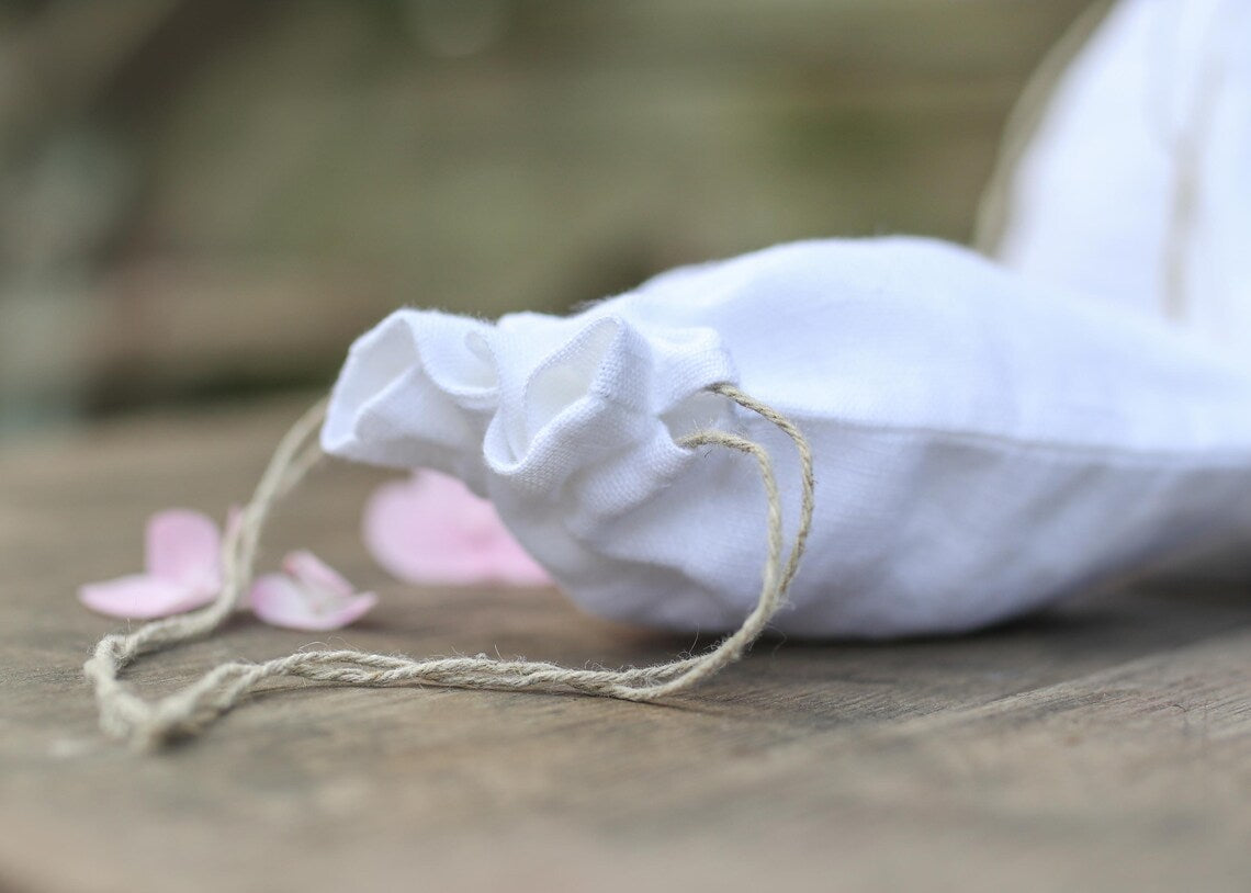 A white linen  fabric drawstring pouch with a natural jute cord, resting on a rustic wooden surface with soft pink flower petals nearby.