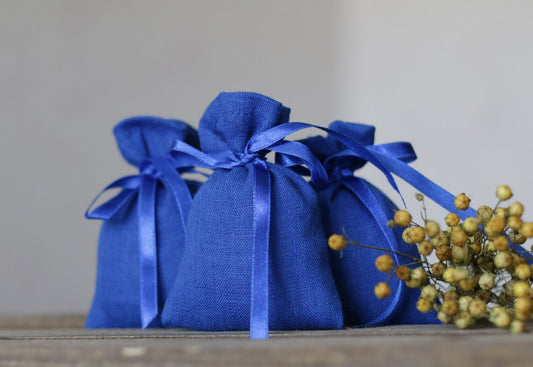Three small blue linen fabric  pouches with matching blue ribbons, arranged on a wooden surface with dried yellow flowers beside them against a soft light gray background.