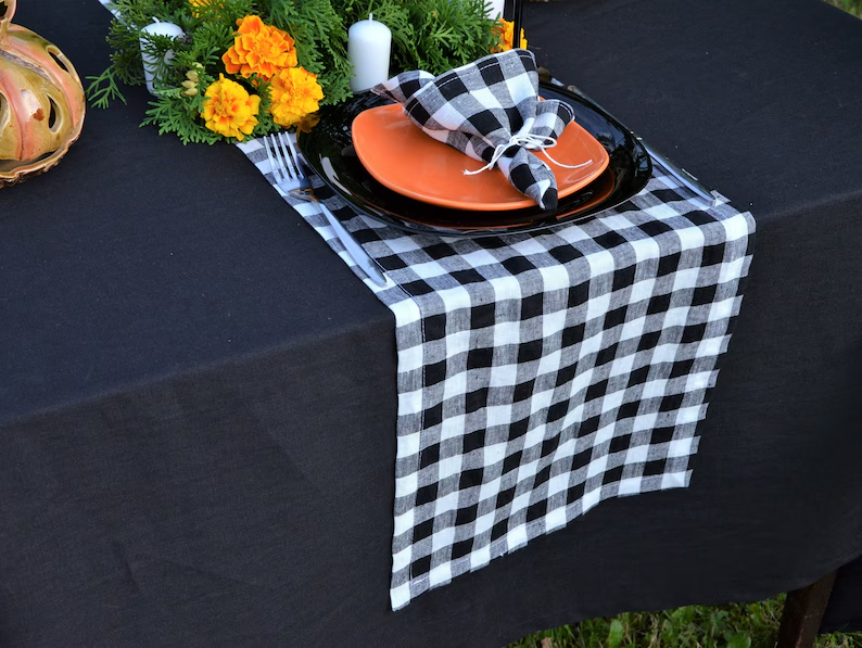 A black and white buffalo plaid table runner draped over a black tablecloth, styled with orange plates, a black napkin, and autumn flowers.