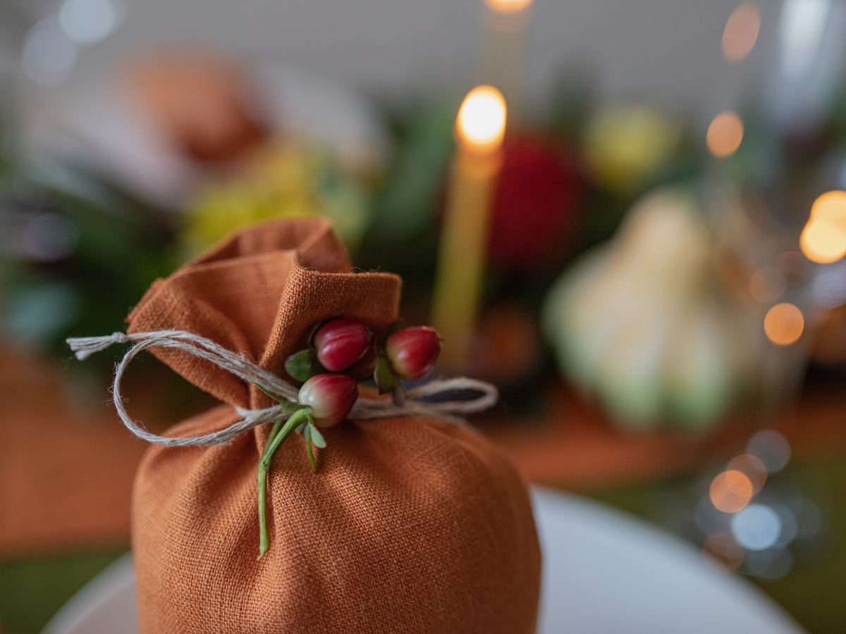 A small rust-orange linen cloth bag tied with twine and decorated with red berry buds, shown in a softly blurred festive tabletop setting with warm lights.