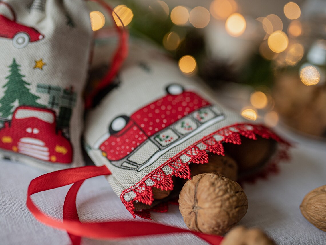 A close-up of a small linen fabric Christmas sack with a red polka-dot truck print, red lace trim, and a red ribbon, with walnuts spilling out and warm blurred holiday lights in the background.