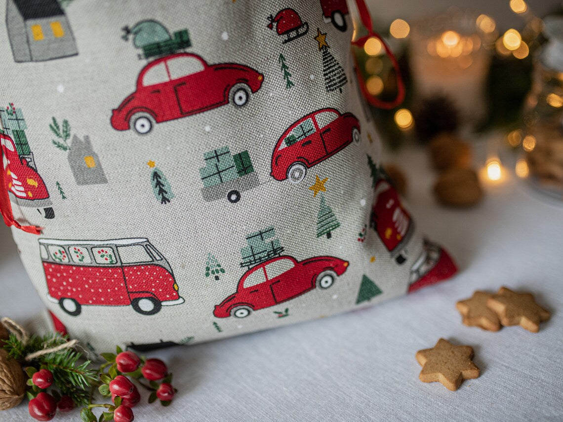 A festive Christmas-themed linen fabric bag or sack with a red vintage car and camper pattern on a light beige background, styled with holiday props like gingerbread cookies, berries, and warm bokeh lights in the background.