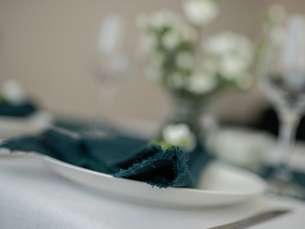 A close-up of a dark teal fringed napkin folded on a white plate, set on a table with a blurred background of glassware and white flowers.