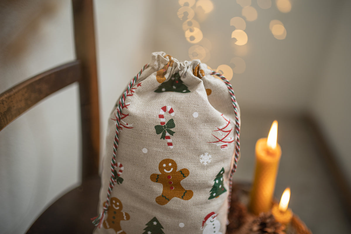A linen fabric Christmas stocking bag with a drawstring closure, printed with gingerbread men, candy canes, and Christmas trees, styled on a wooden chair next to lit candles.
