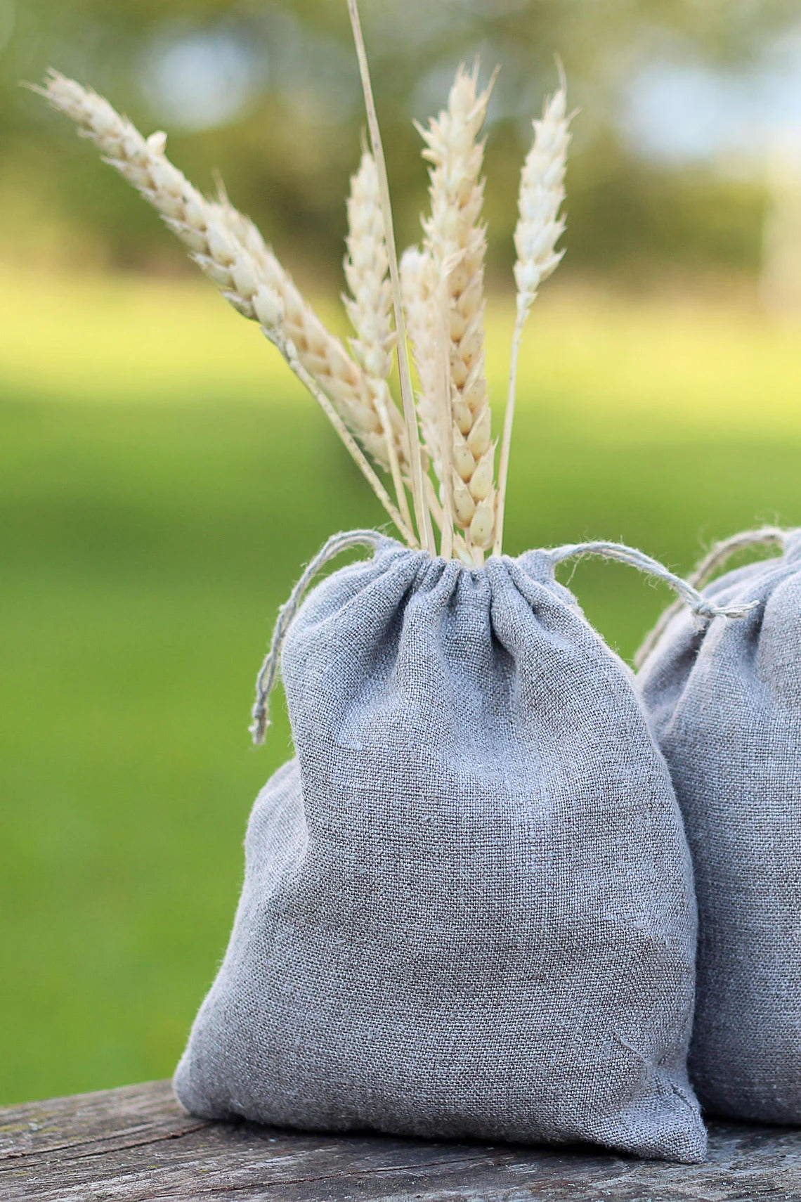 A small gray (not dyed) linen drawstring bag filled with dried wheat stalks, sitting on a wooden surface with a blurred green outdoor background.