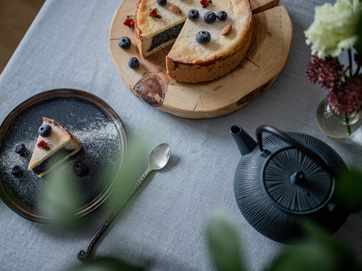 Cheesecake with berries on a wooden board, slice on a plate, teapot, and flowers on a dusty blue linen tablecloth
