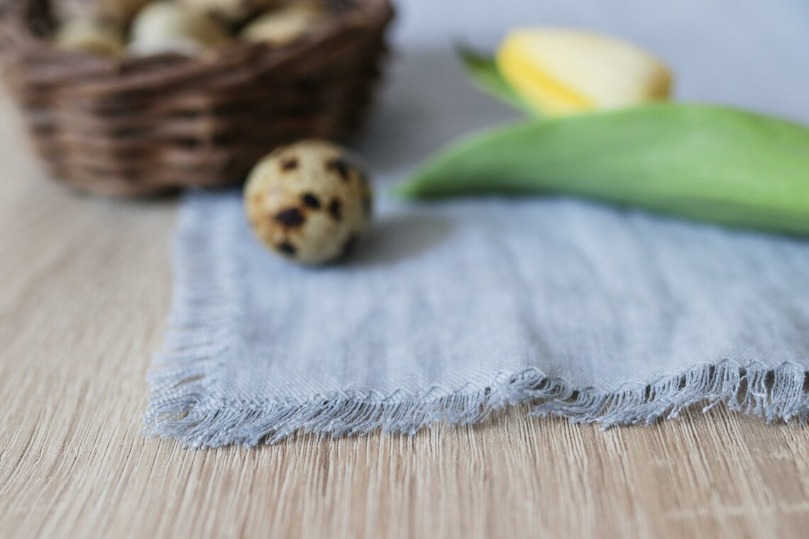 A close-up of a light blue fringed linen table runner on a light wood surface, with a speckled quail egg, a wicker basket, and a yellow tulip in soft focus in the background.