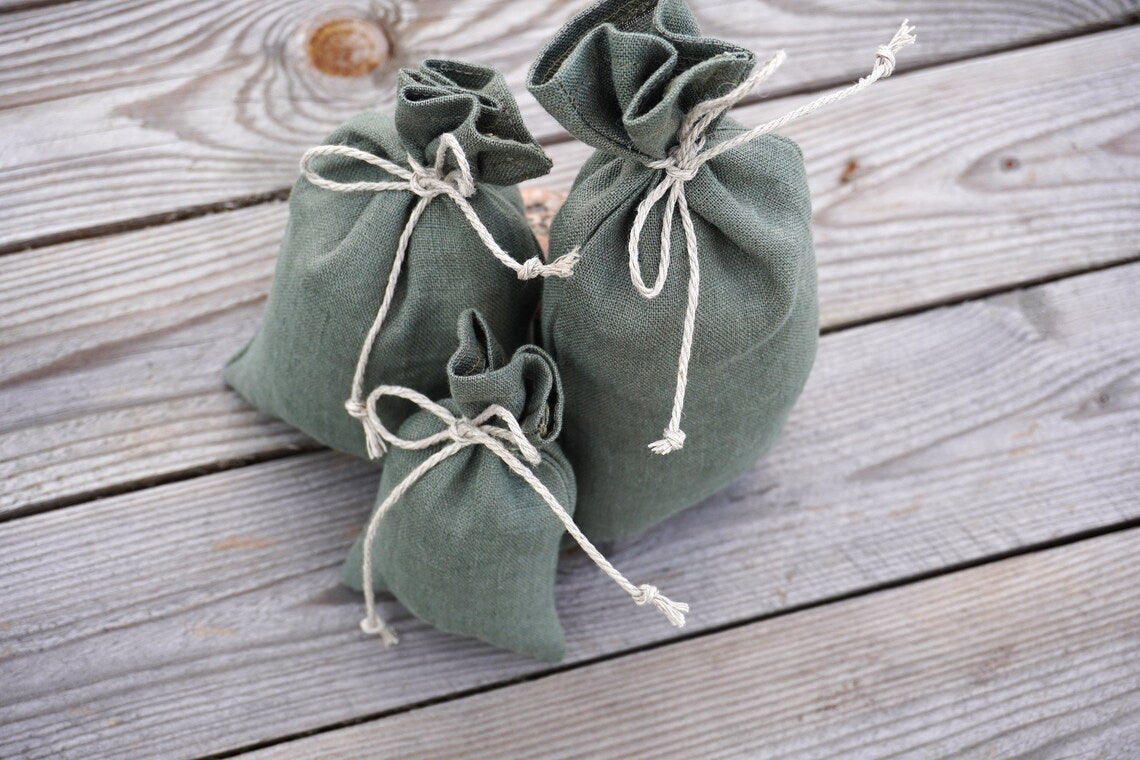 Three small sage green linen gift bags  made of textured fabric, tied with white rope, arranged on a rustic gray wooden plank surface.