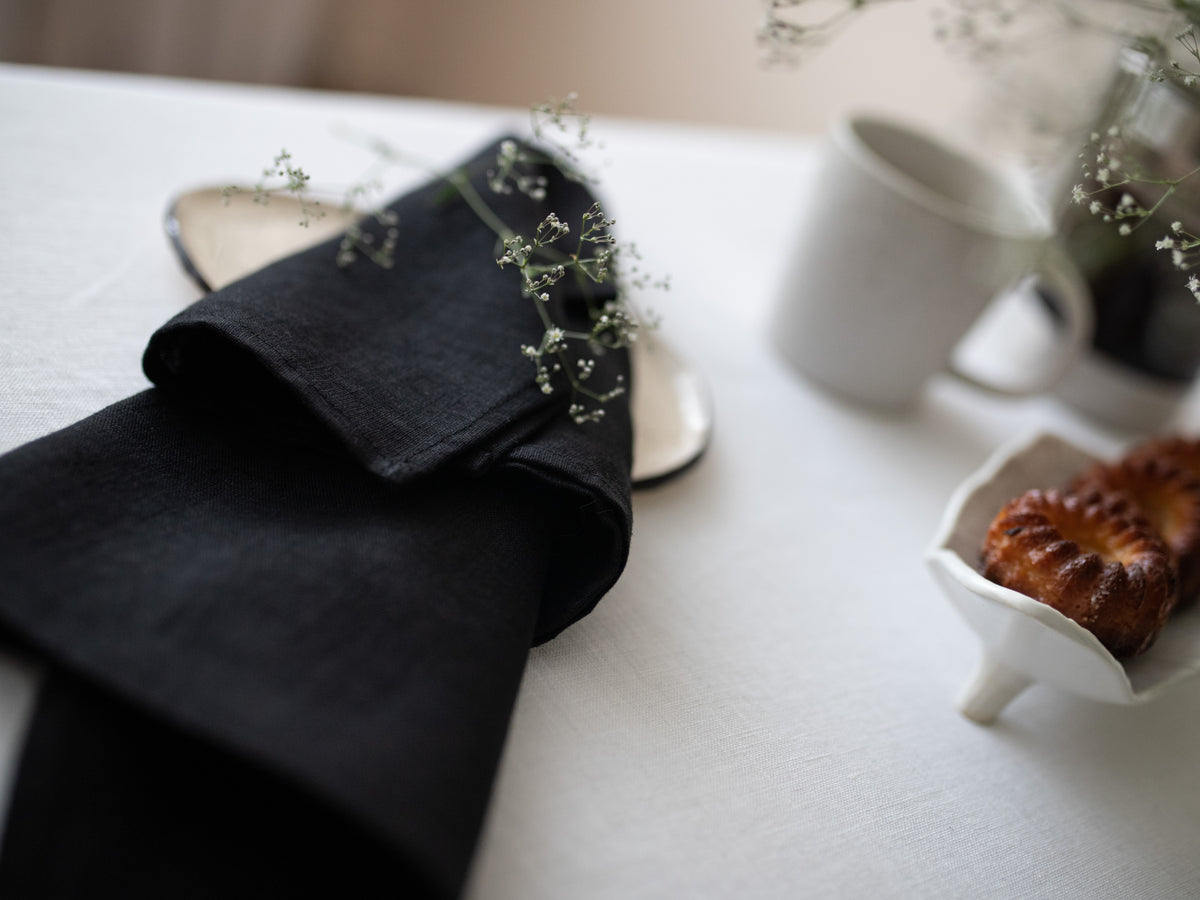 A folded black linen napkin with delicate baby's breath flowers on top, styled on a white tablecloth alongside a white ceramic mug and a small pastry dish.