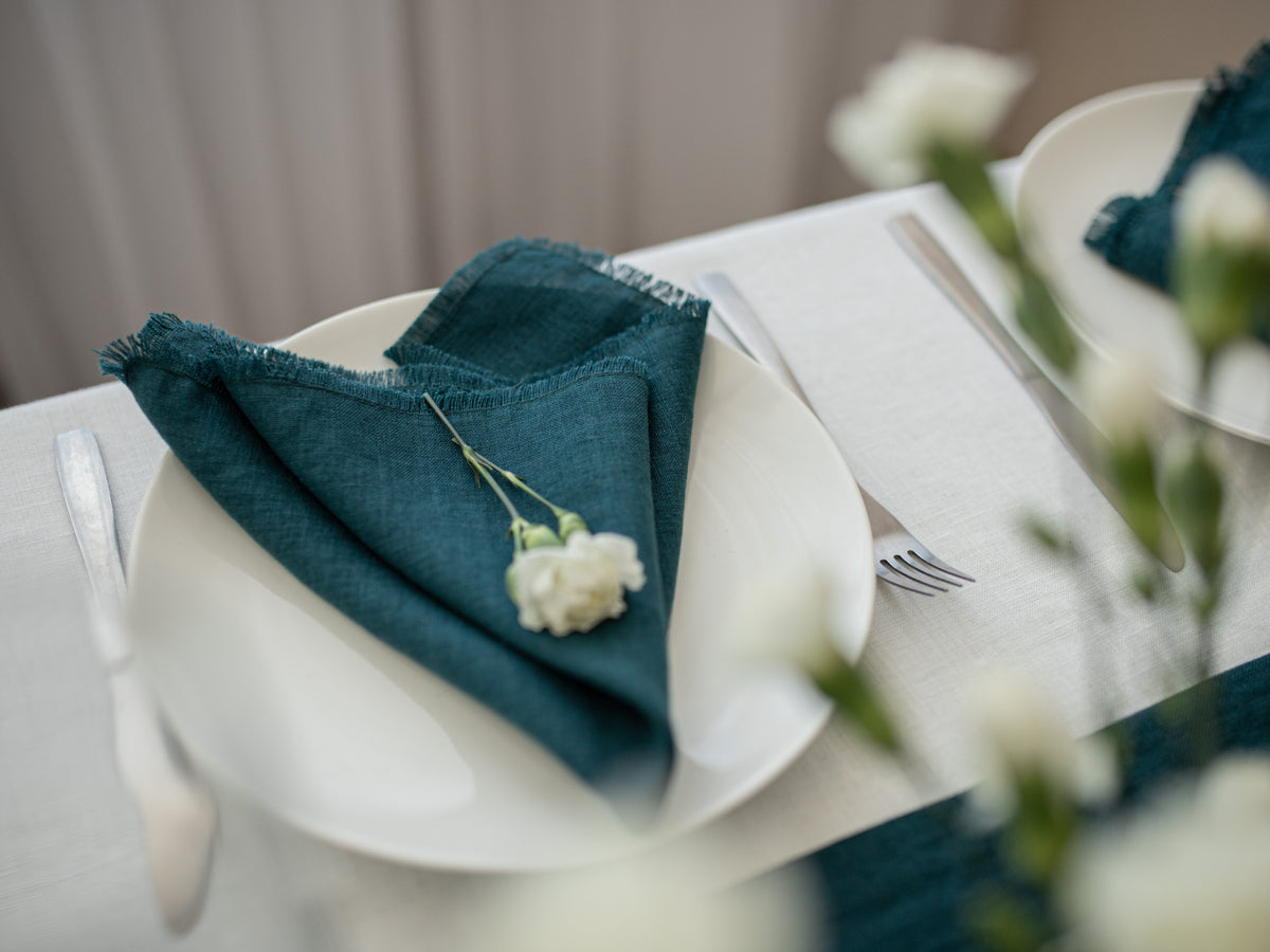A styled table setting featuring a dark emerald green linen napkin with frayed edges, folded over a white plate and adorned with a small white flower. Silver flatware and a matching blurred napkin are visible on a white tablecloth.