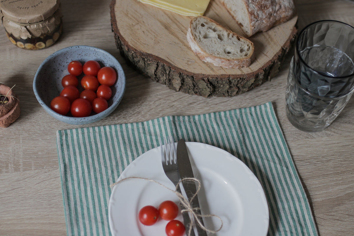 A styled tabletop setting with a white plate, silver fork and knife tied together with twine and topped with a cherry tomato, placed on a green and white striped napkin. A blue bowl of cherry tomatoes, a rustic wood slice with bread, and a textured glass are arranged around it on a light wood table.
