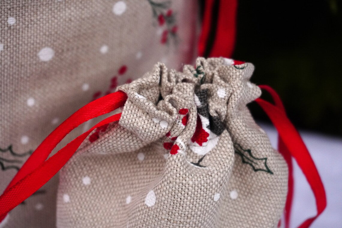 A close-up of a beige burlap drawstring bag with white snowflake patterns, robin,  red ribbon ties, and small holly leaf prints.