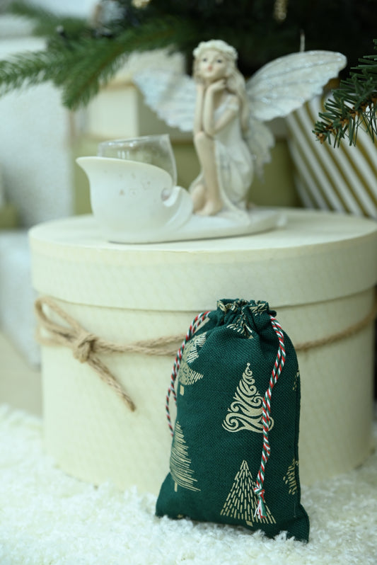 A dark green Christmas-themed drawstring pouch with a gold tree pattern and red-and-white striped cord, placed in front of a cream-colored gift box with twine, a white angel figurine, and evergreen branches.