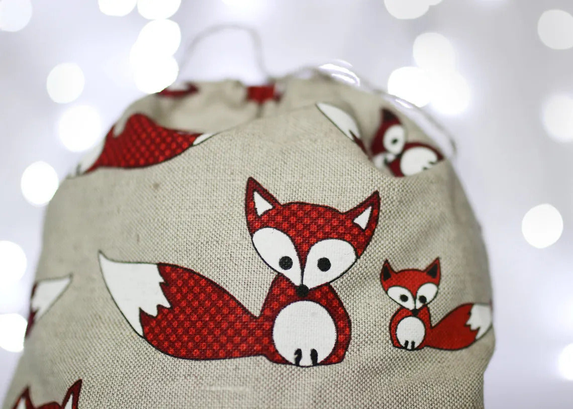 A close-up of a fabric drawstring bag featuring a red and white fox pattern on a beige linen-like background, with a soft bokeh light background.