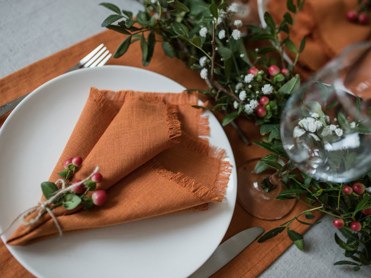 Table setting with white plate,  orange fringed napkin, and greenery on a wooden table.