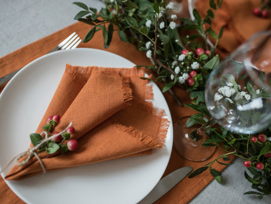 Table setting with white plate,  orange fringed napkin, and greenery on a wooden table.
