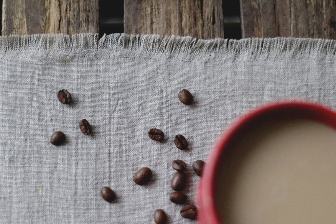 Dark roasted coffee beans scattered on a not dyed  linen table runner, with a red cup of coffee partially visible in the foreground on a rustic wooden table.