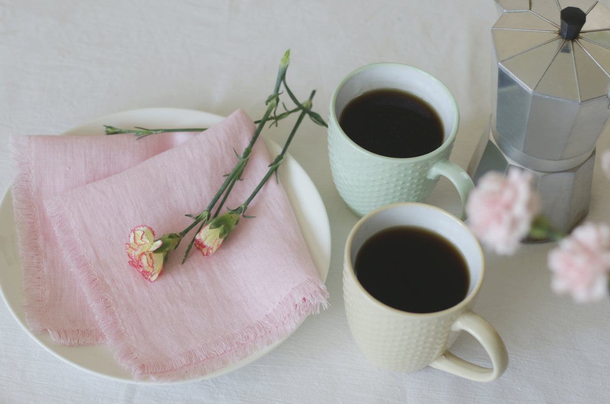 Two cups of coffee on a table with a pink cloth linen napkin and flowers.