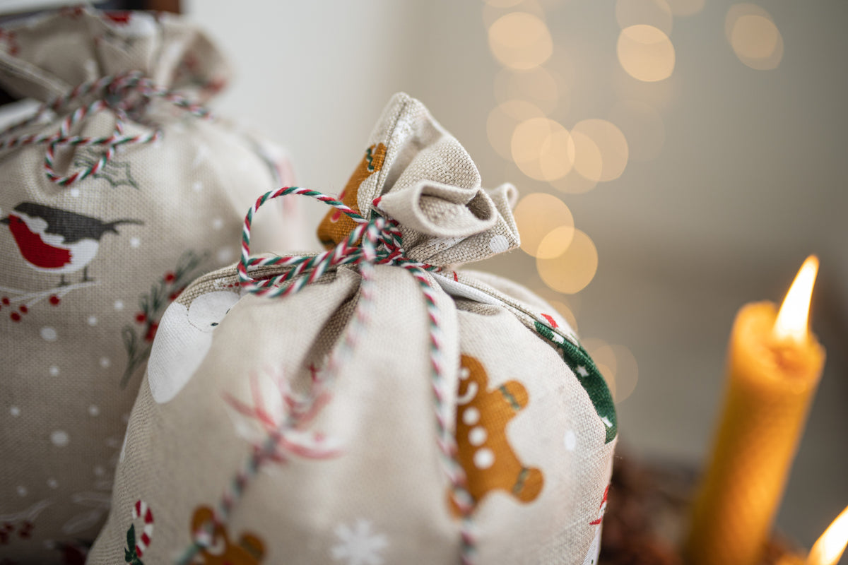 A close-up of a linen fabric gift bag with a holiday print featuring gingerbread cookies, snowflakes,  tied with red, white, and green bakers twine. A lit yellow candle glows in the warm, blurred background.