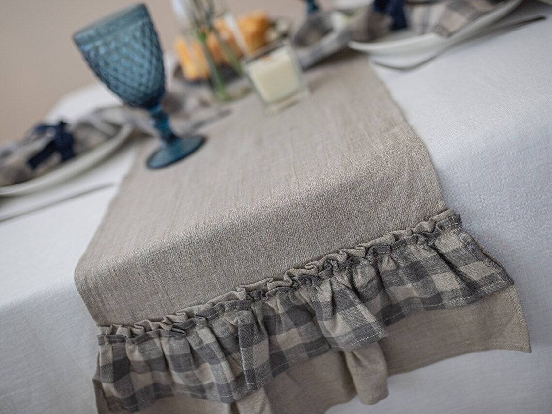 A not dyed linen table runner with a gathered ruffle trim in a gray and beige plaid pattern, styled on a white tablecloth with dining tableware in the background.