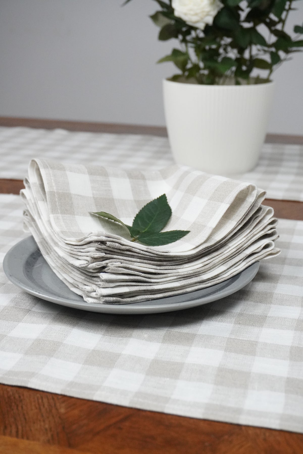 Stack of folded plaid linen napkins on a wooden surface with a checkered tablecloth and a vase with flowers.