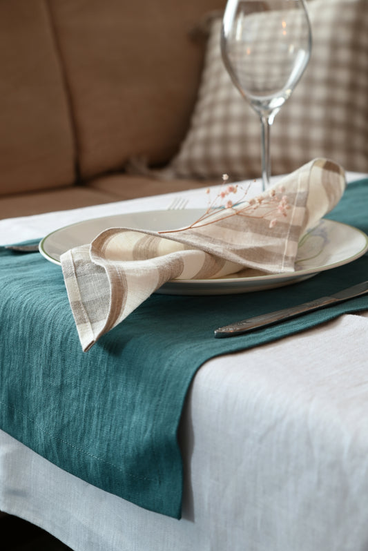 Dining table setting with a plate, folded striped linen napkin, and wine glass on a green runner.