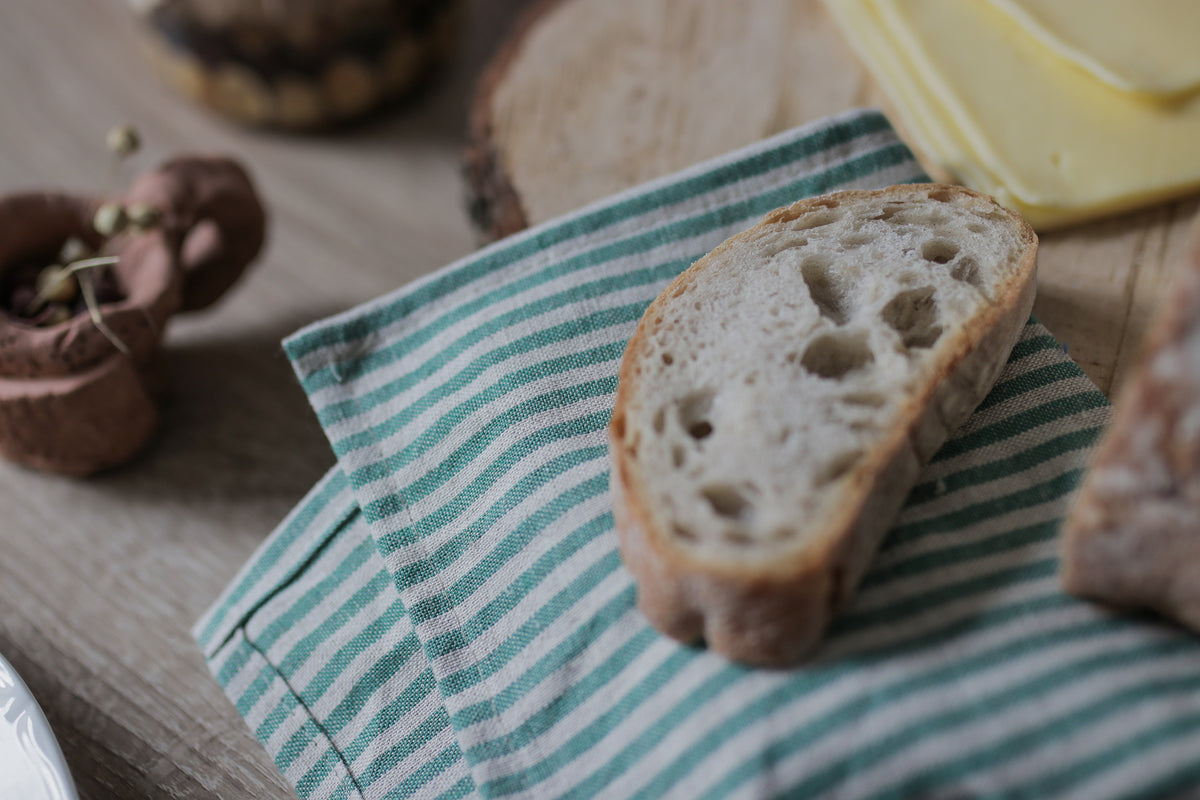 A rustic slice of artisanal bread with a green and white striped cloth linen  napkin underneath, set on a wooden table with cheese and other food items blurred in the background.