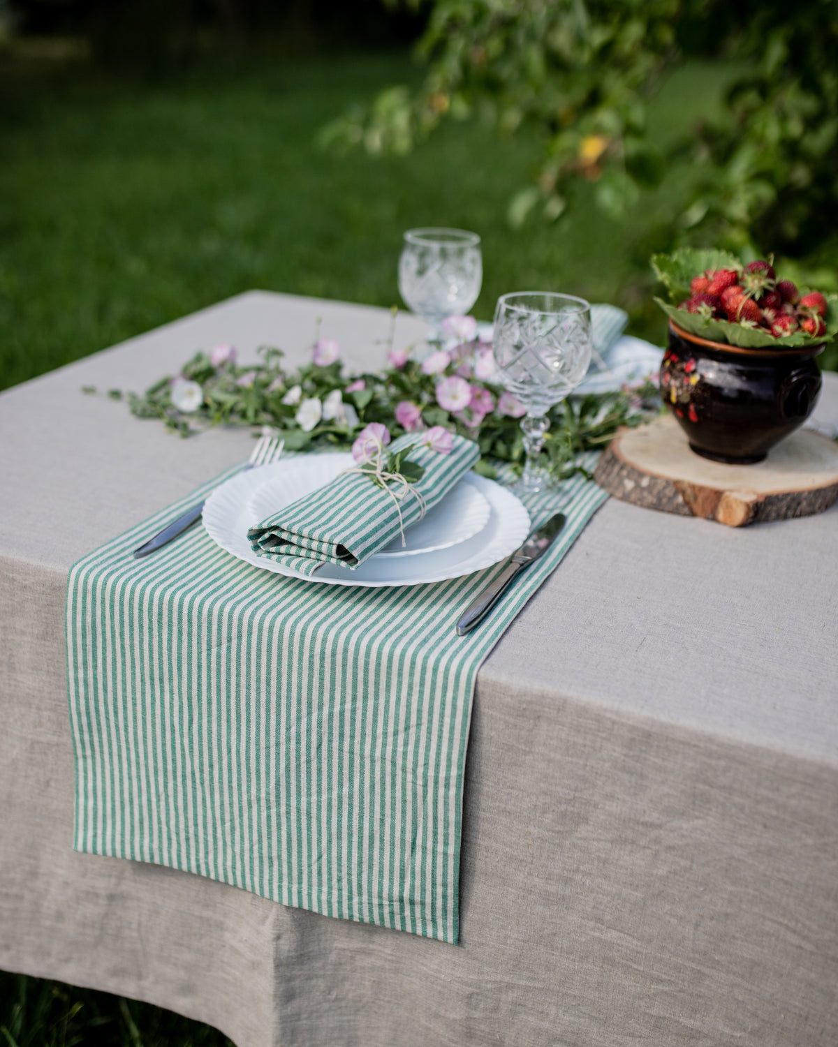 Outdoor table setting with green and white stripped linen runner, plates, glasses, and flowers.