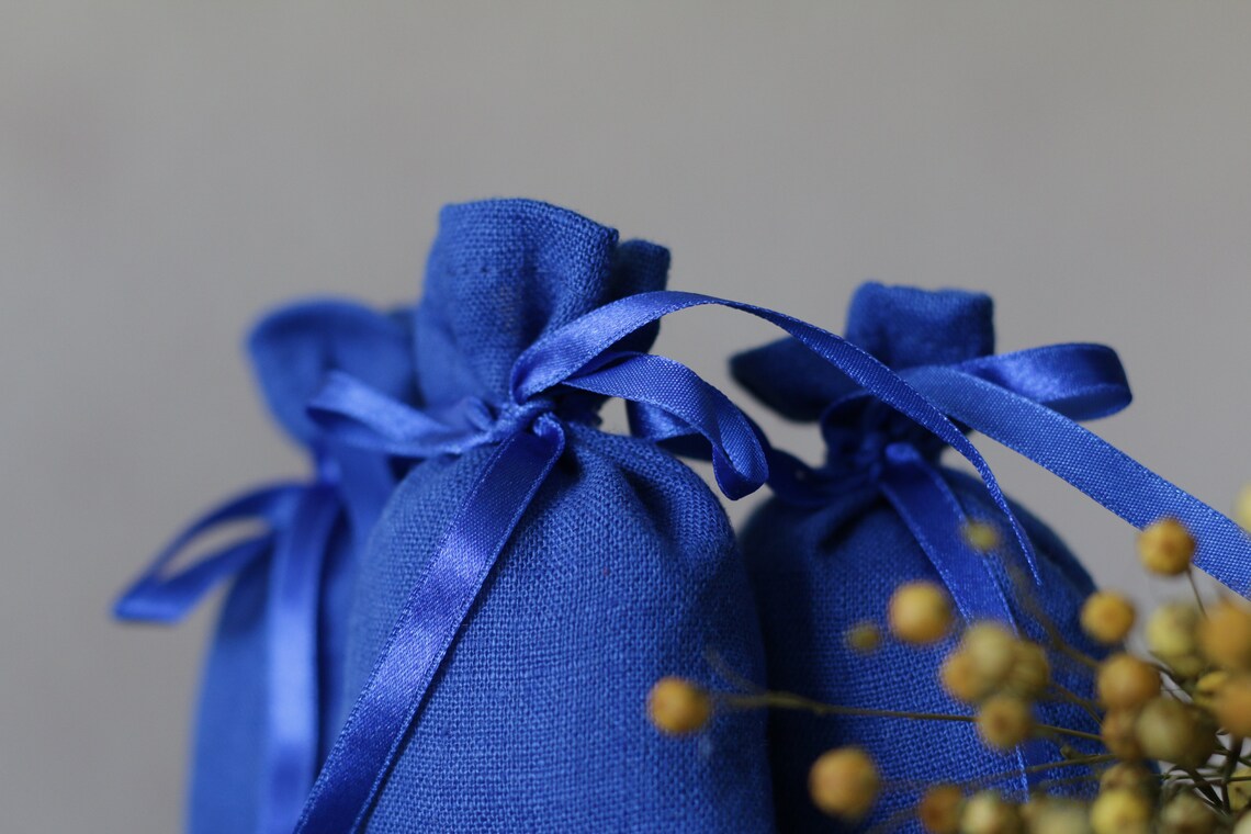 Three linen royal blue  linen pouches with royal blue satin ribbons, arranged with small yellow dried flowers against a soft gray background. 