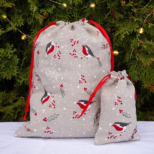 Two linen fabric drawstring gift bags with a robin bird and holly berry pattern, displayed in front of a decorated Christmas tree.