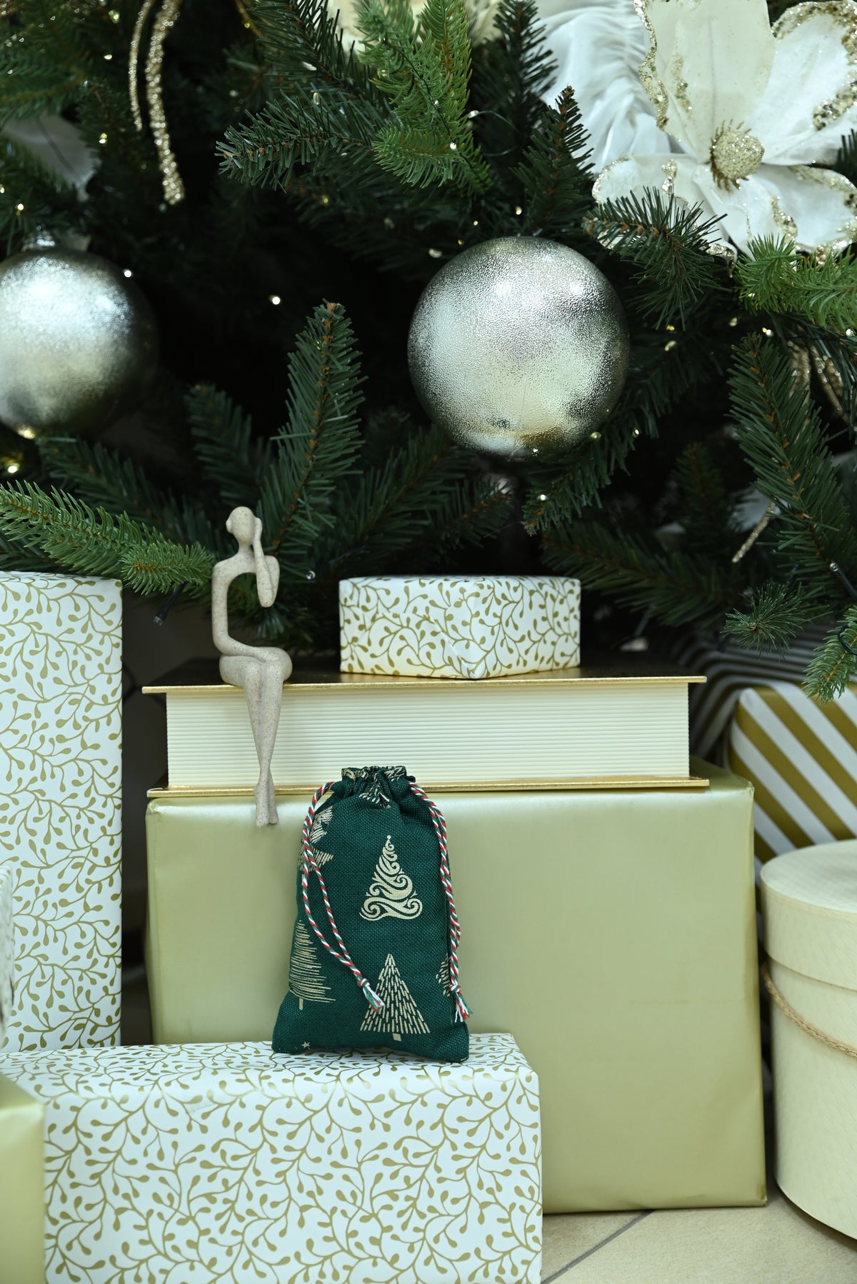 A dark green drawstring gift pouch with a white Christmas tree pattern sits on top of wrapped gift boxes in front of a decorated Christmas tree with silver ornaments.