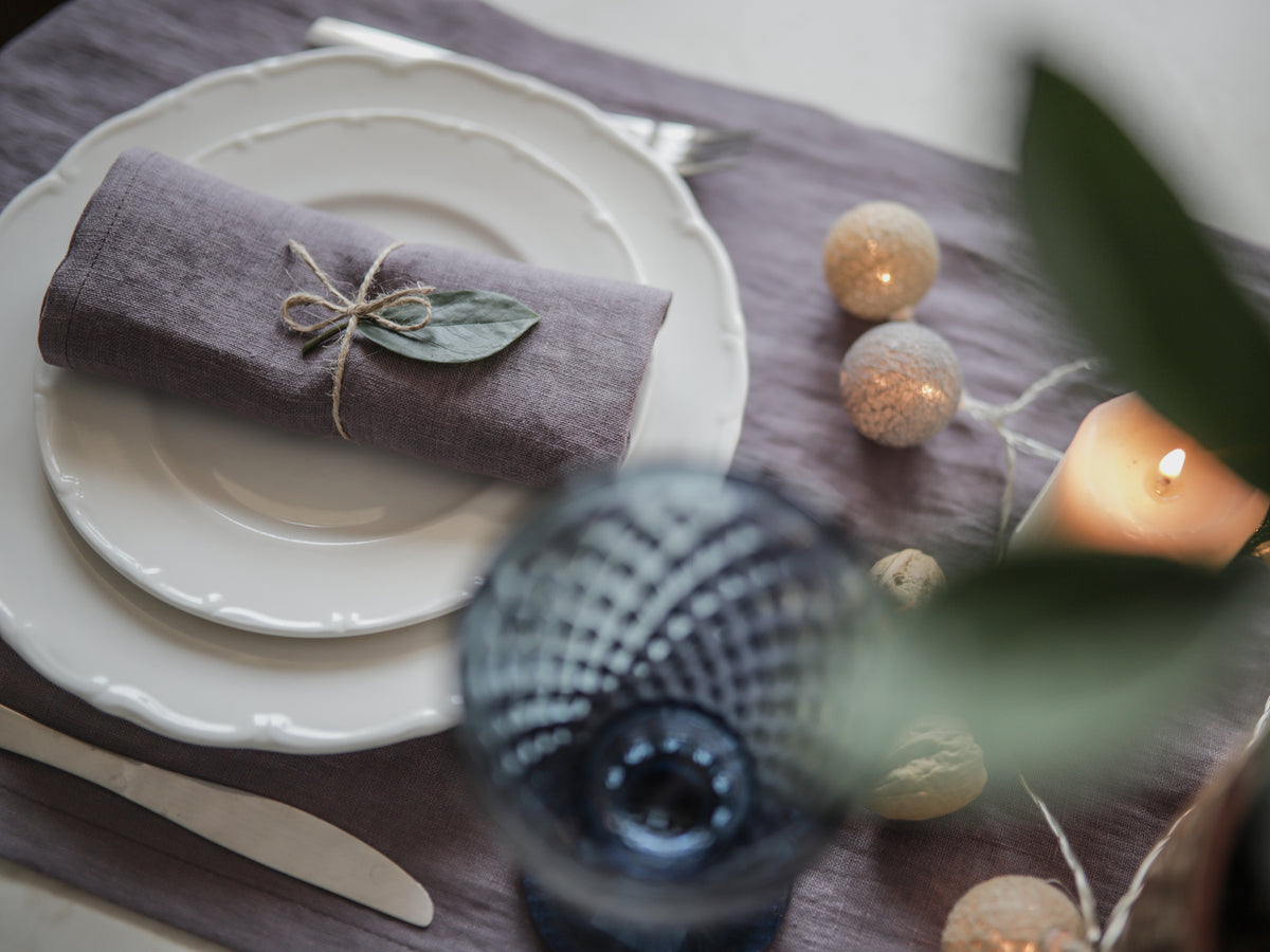 A styled table setting featuring a folded dusty purple linen napkin tied with twine and a small green leaf, placed on a white plate. The scene includes a purple table runner, a blue textured glass, a lit candle, and warm decorative lights.
