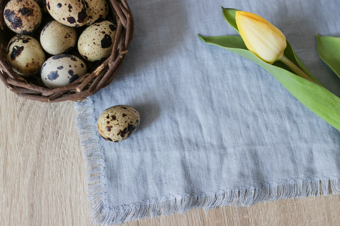 A styled flat-lay featuring a dusty blue linen table runner with fringed edges, a brown woven basket of speckled quail eggs, and a yellow tulip on a light wood surface.
