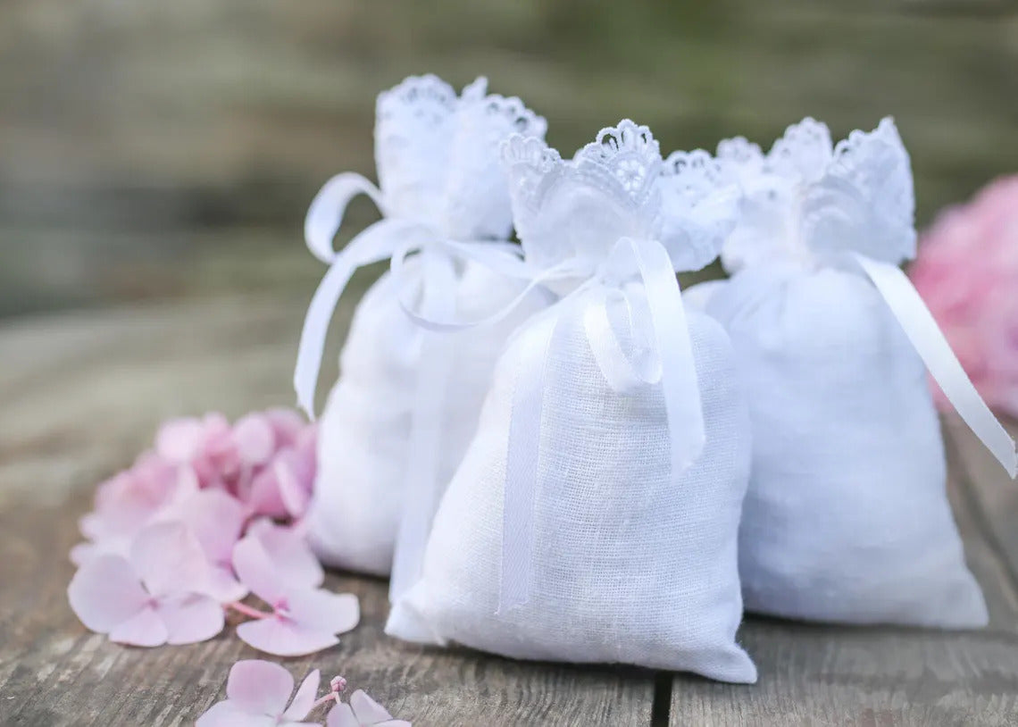 Three small white linen fabric bags with lace trim and white ribbons, arranged on a rustic wooden surface with pink flower petals around them.
