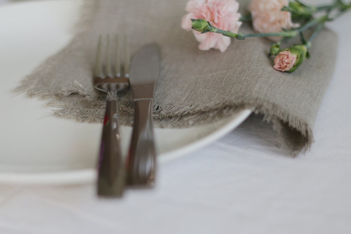 Silverware on a white plate with a nor dyed  fringed linen napkin and pink flowers.