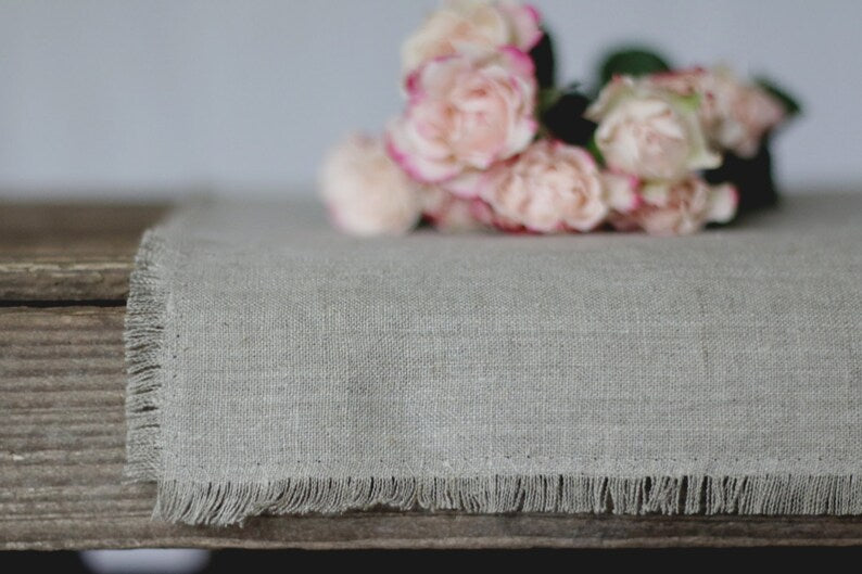 A light gray linen table runner with fringed edges draped over a rustic wooden table, styled with a bouquet of soft pink roses in the background.
