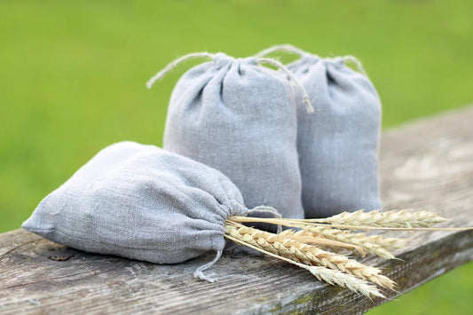 Three small gray linen drawstring pouches arranged on a weathered wooden surface with wheat stalks in front, against a blurred green outdoor background.
