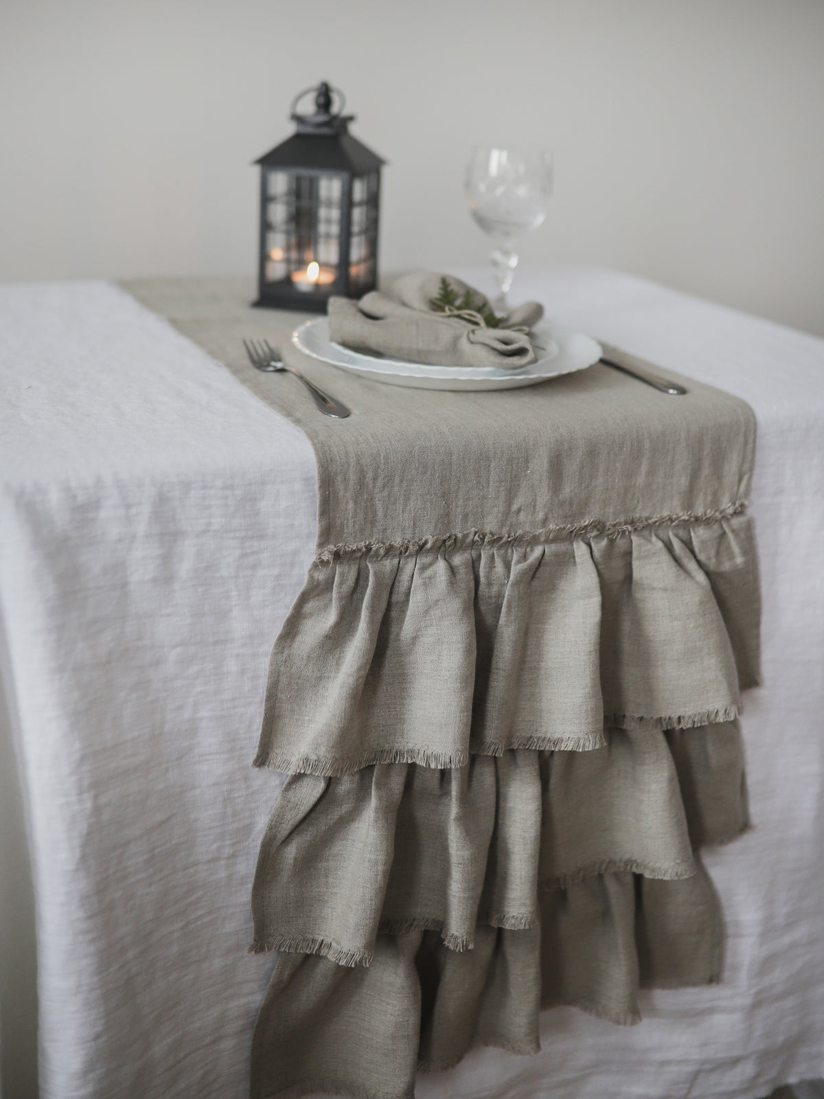 A natural color linen table runner with tiered ruffles and frayed edges, styled on a white tablecloth-covered table with a white plate, folded napkin, glass, and a black lantern.