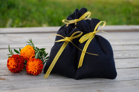 Two small navy blue  linen fabric pouches tied with shiny gold ribbons, placed on a weathered wooden surface outdoors with orange marigold flowers beside them.