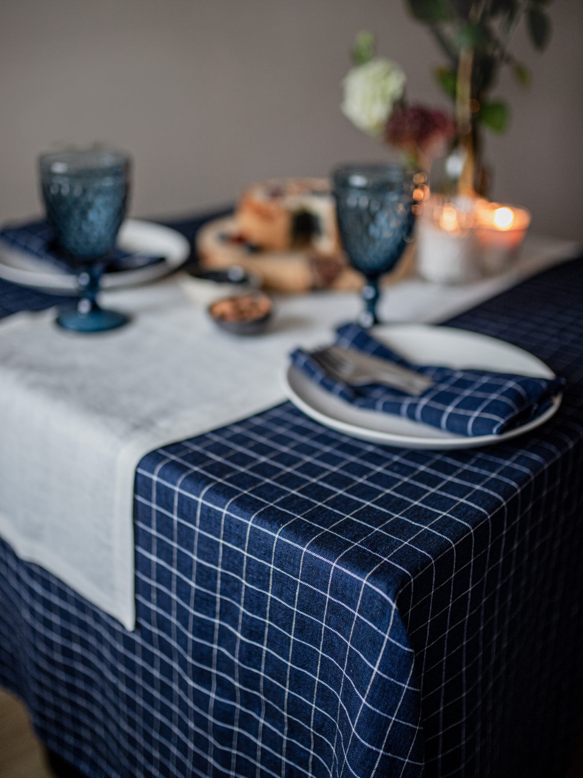 Dining table setting with blue checkered linen tablecloth, white plates, and blue glasses.