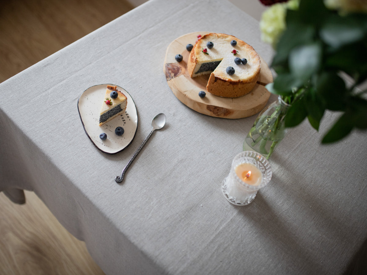 Dessert setting with a slice of cake on a plate, a whole cake on a wooden board, a candle, and a spoon on a natural linen tablecloth.