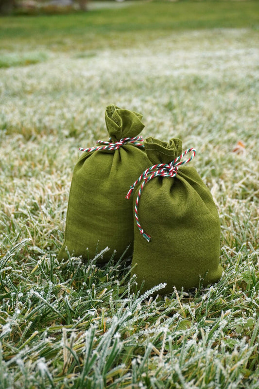 Two olive green linen drawstring pouches tied with red, white, and blue striped twine, standing on frosty grass outdoors.