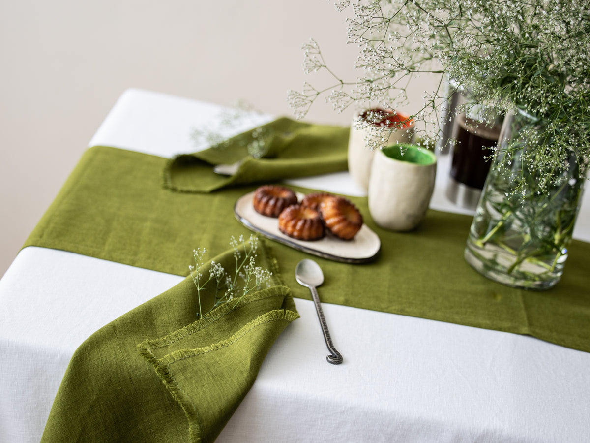 Table setting with green runner, olive green linen napkin, white tablecloth, and small dessert plate.