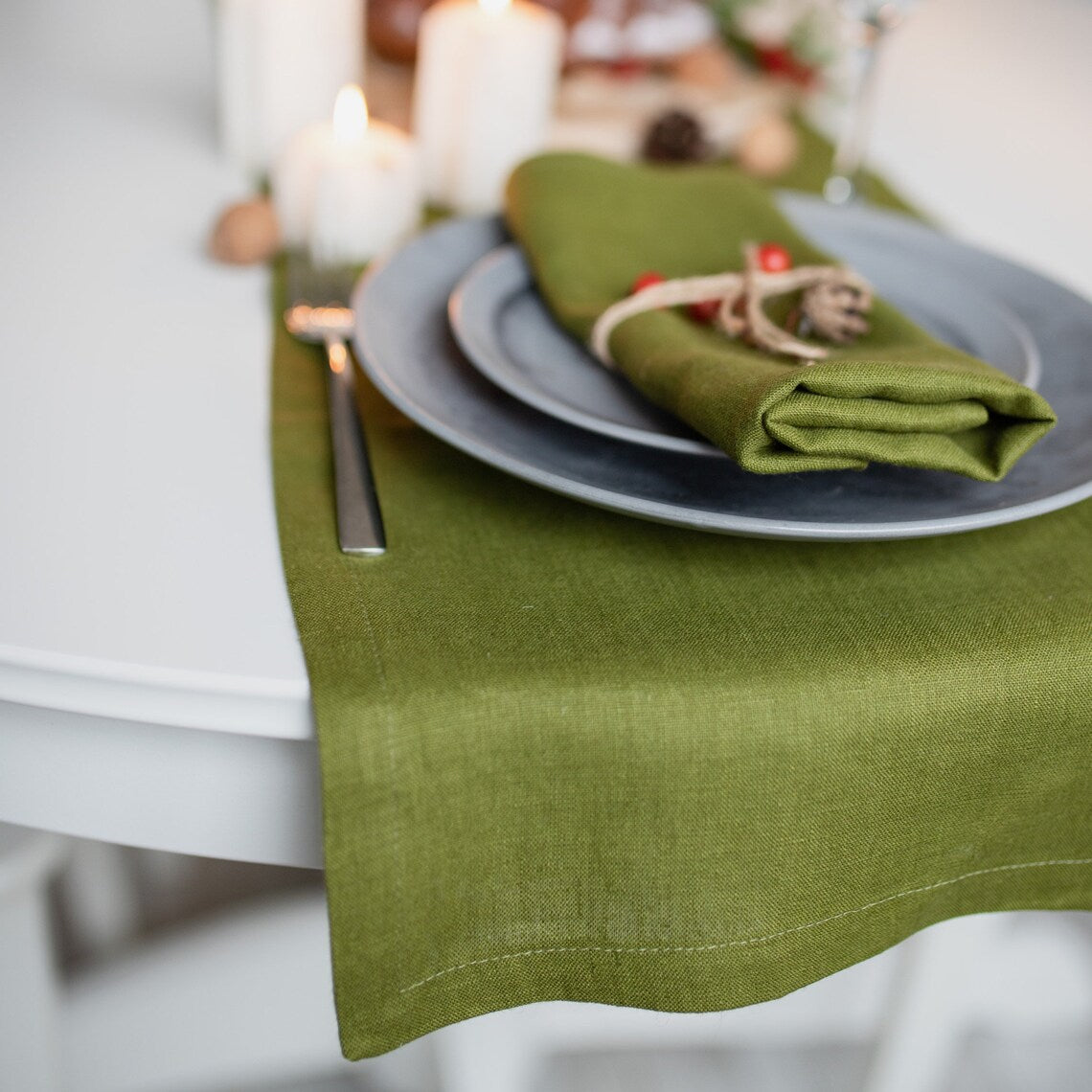 An olive green linen table runner draped over a white table, styled with a matching folded napkin on a gray plate and a silver fork.