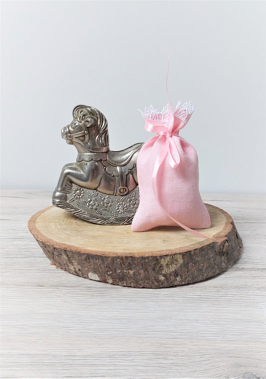 A small light pink linen pouch with white lace trim sits on a round natural wood slice coaster next to a silver rocking horse figurine, set against a light gray background.