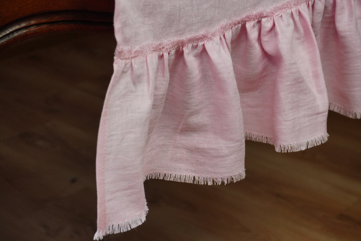 A close-up of a light pink linen table runner with frayed edges and a ruffled hem, draped over a wooden table