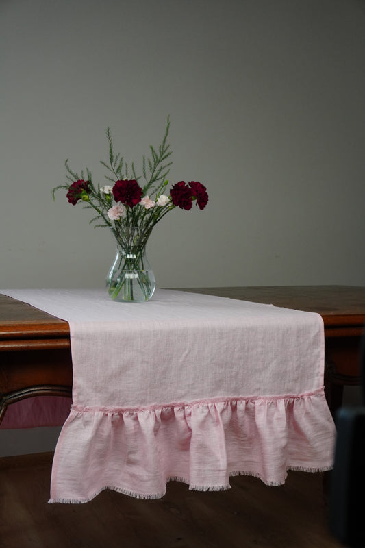 A light pink linen table runner with a ruffled hem is draped over a dark wooden table, styled with a clear glass vase containing dark red and white flowers against a plain gray wall.