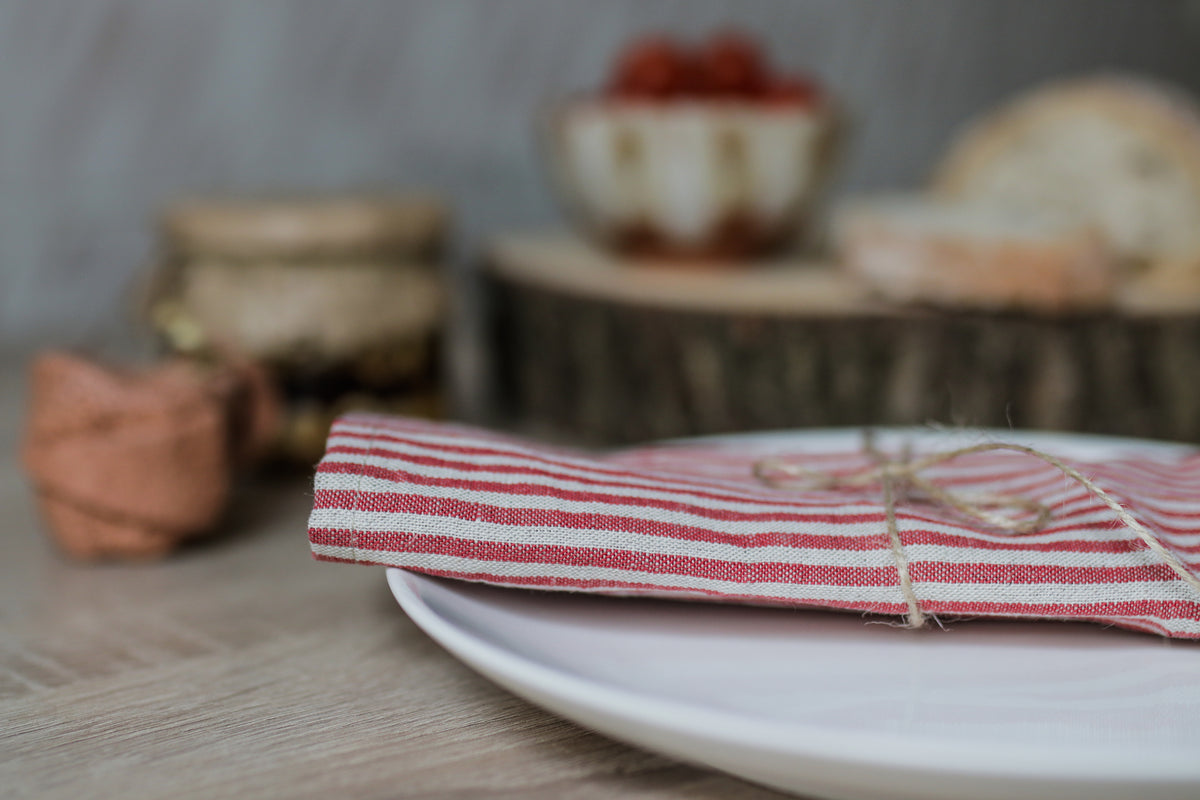 A folded red and white striped linen napkin tied with twine, resting on a white plate on a light wood table, with a softly blurred rustic background.